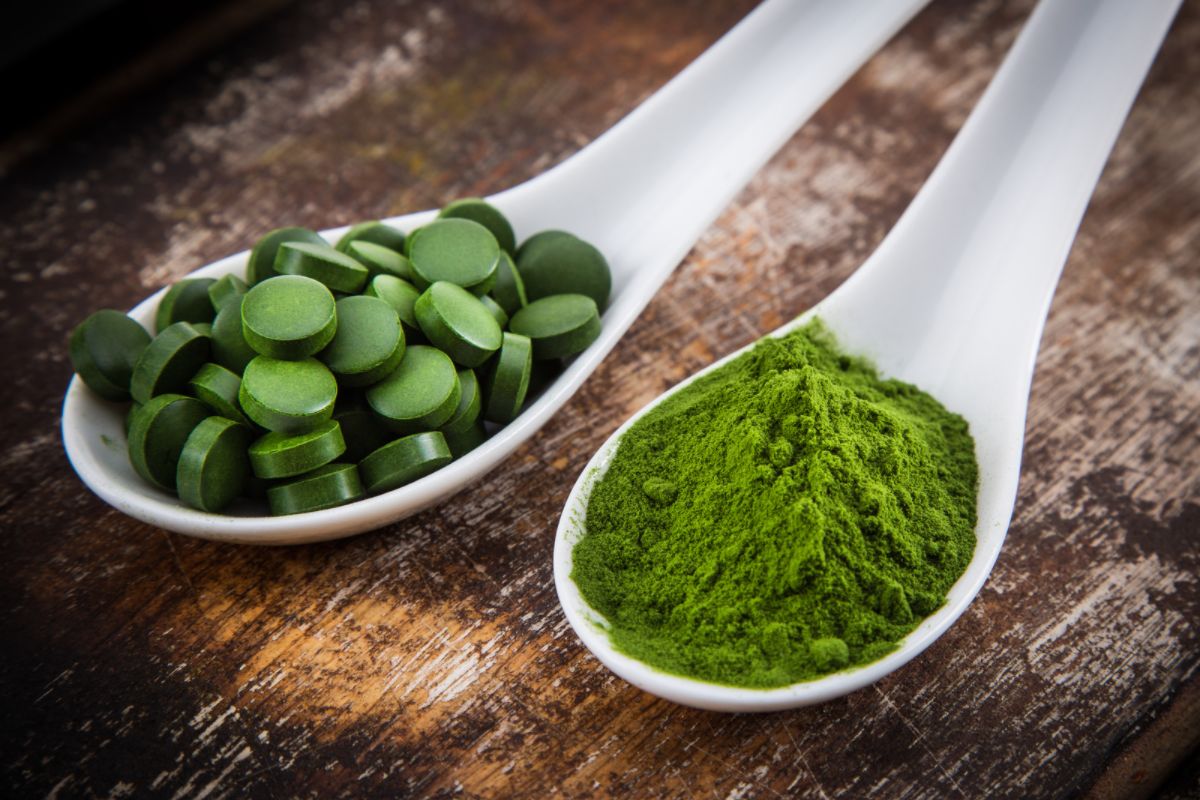 One ceramic spoon with chlorella powder and another with tablets are shown closeup on a wooden tabletop