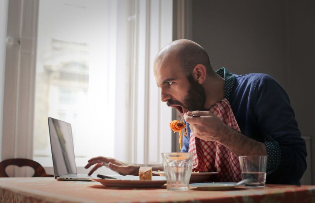 A man sitting at a dining room table, jaw dropped, with a fork full of spaghetti in one hand and scrolling on his laptop with the other as he stares at the screen.