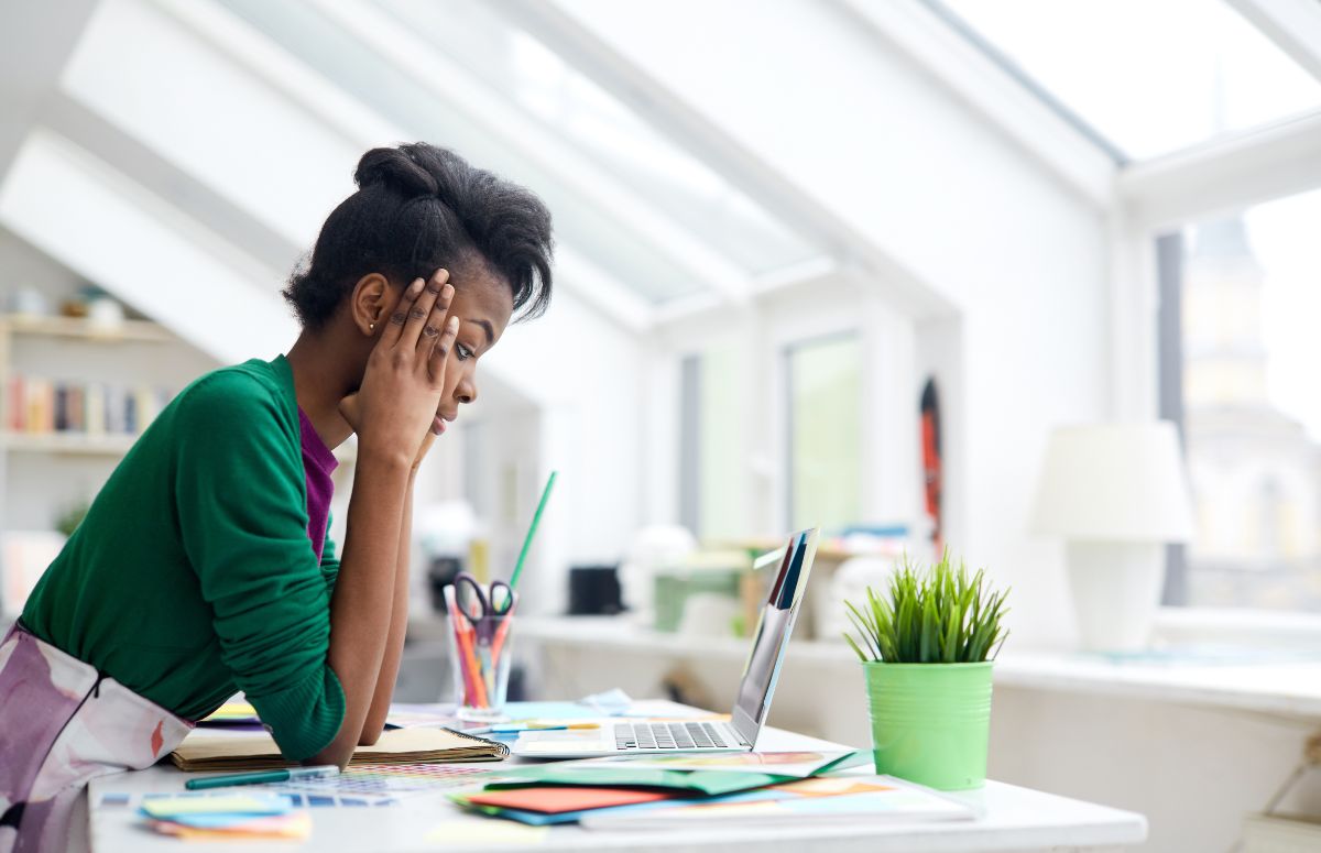 A distraught woman with hands pressed to her temples peers down at her laptop on a desk filled with documents and folders