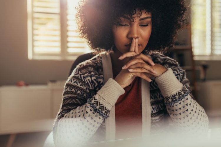 A woman sits at a desk with eyes closed and clasped hands over her lips in deep reflection